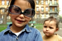Young girl in a blue shirt wearing upside-down glasses. A small child is behind looking over her shoulder and block of flats is in the background