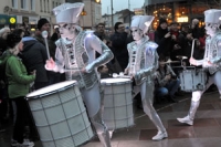 Performance artists dressed in white costumes and hats, with white painted faces, playing drums in front of an animated crowd