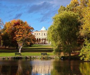A view of Pittville Pump Room across the lake and through the trees in Pittville Park. The Pump Room is a Regency building fronted by tall columns and topped with a domed roof.