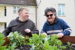 Two people smiling and leaning on a wooden planter containing veg plants