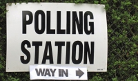 Sign which reads 'polling station - way in' attached to a wire fence with green hedge behind
