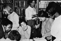 Young white and Caribbean women smiling and looking at books in a library. Black and white photo from around 1960.